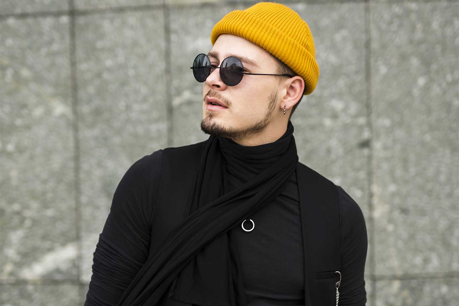 Hombre joven con gorro amarillo y gafas de sol negras, luciendo un estilo moderno y elegante con prendas negras. Fondo de pared de piedra gris.